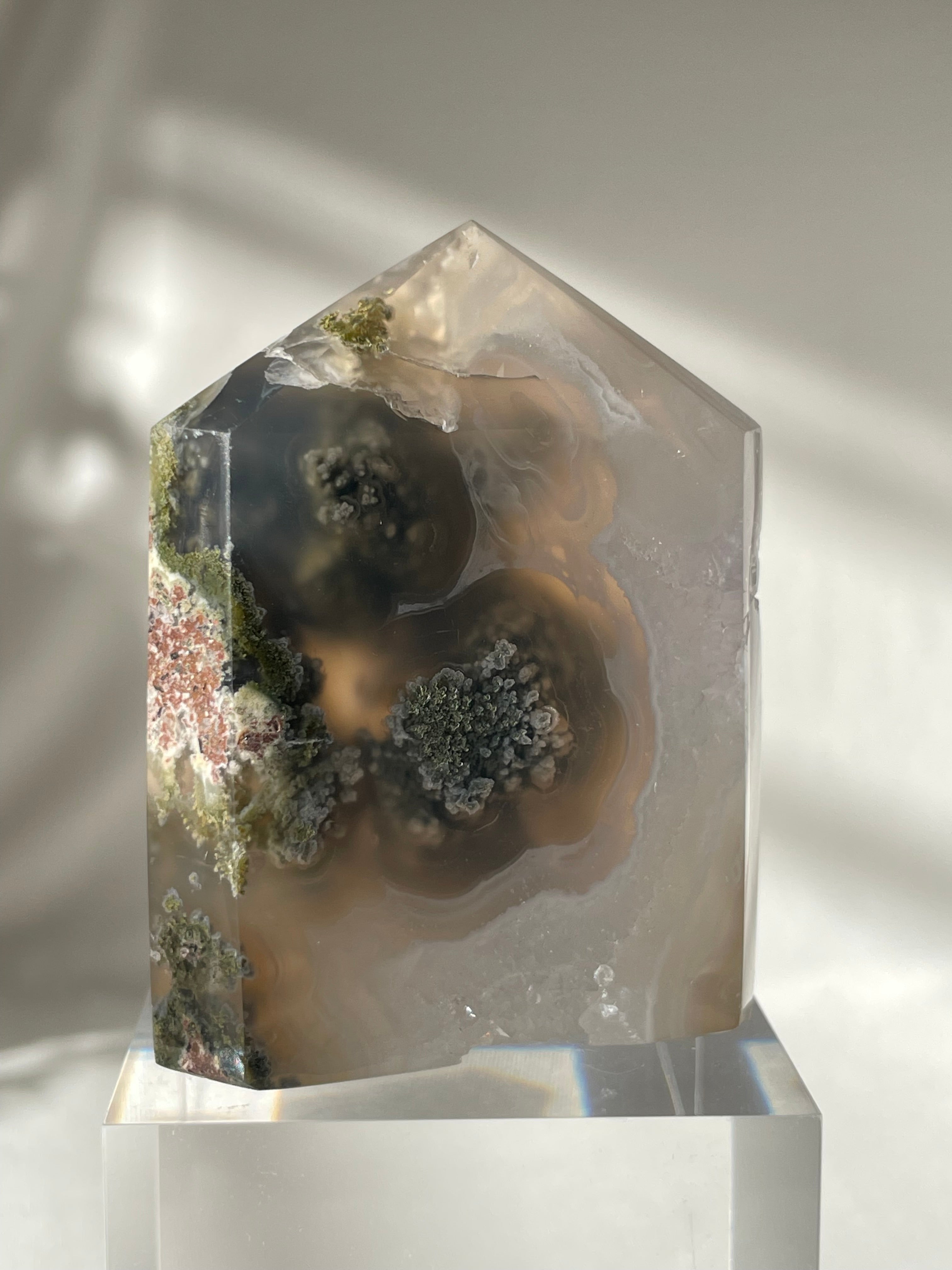Scenic moss agate crystal tower with intricate patterns and earthy tones, displayed on a clear pedestal.