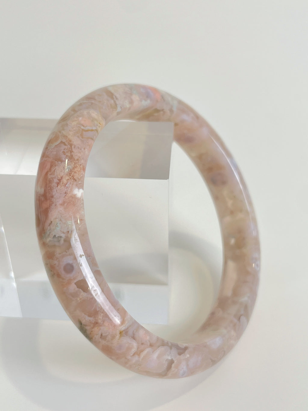 Flower agate bangle with soft pink hues, displayed against a clear stand, showcasing its smooth surface.