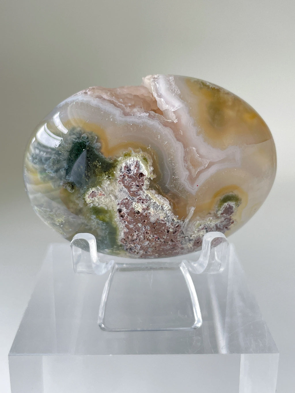 Scenic moss agate palm stone with earthy tones displayed on a clear stand, showcasing its unique patterns.