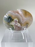 Scenic moss agate palm stone with earthy tones displayed on a clear stand, showcasing its unique patterns.