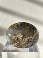 Scenic moss agate palm stone with earthy tones and unique patterns, resting on a clear display stand.