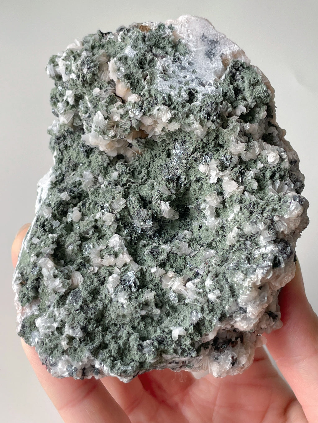 A hand holding a textured green and white mineral specimen, showcasing intricate crystal formations.