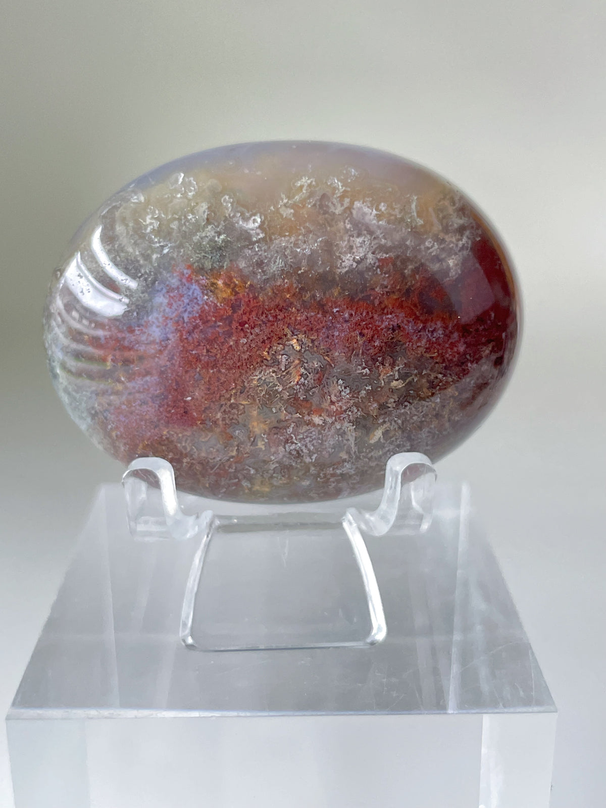 Scenic moss agate palm stone with rich earthy tones displayed on a clear stand, showcasing its natural beauty.