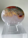 Scenic moss agate palm stone displayed on a clear stand, showcasing vibrant colors and unique patterns.