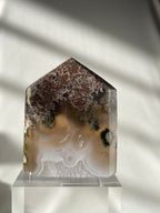 Scenic moss agate tower with earthy tones and unique patterns, displayed on a clear stand against soft lighting.