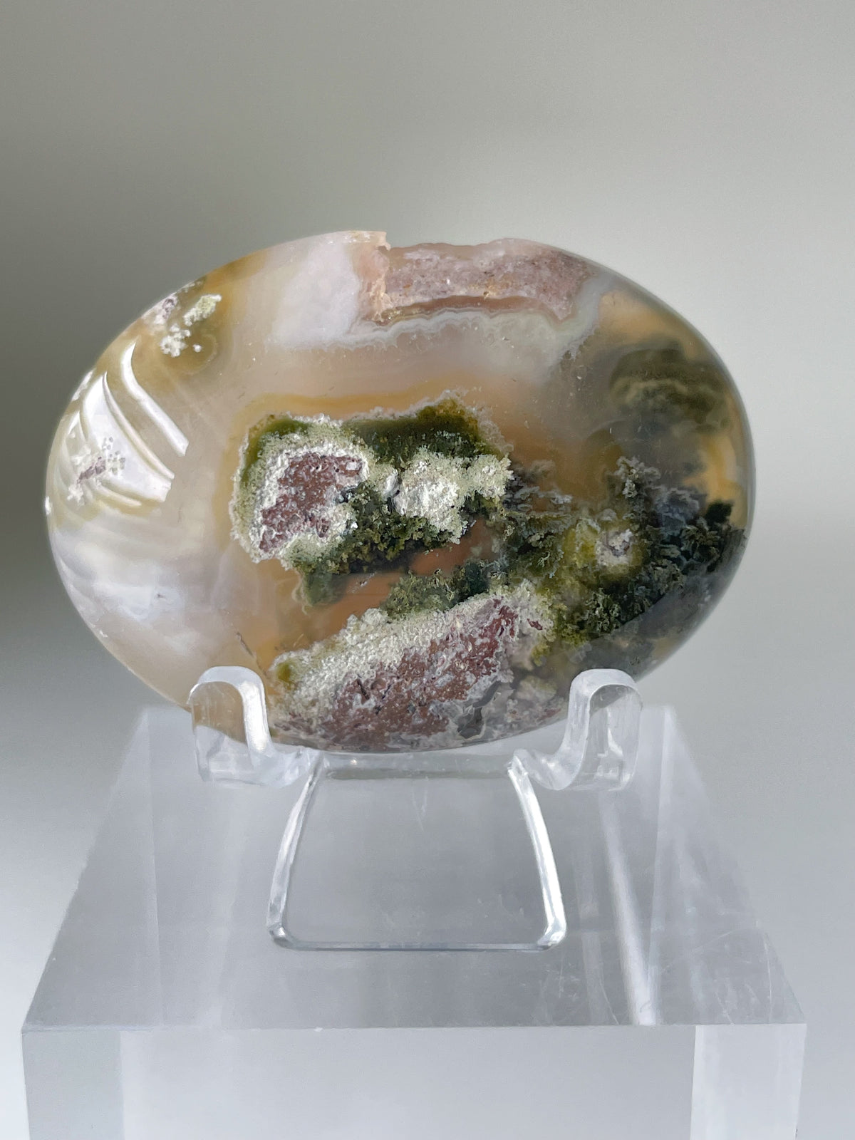 Scenic moss agate palm stone displayed on a clear stand, showcasing earthy tones and intricate patterns.