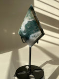 Scenic moss agate crystal specimen displayed on a sleek black stand, showcasing its unique green and white patterns.