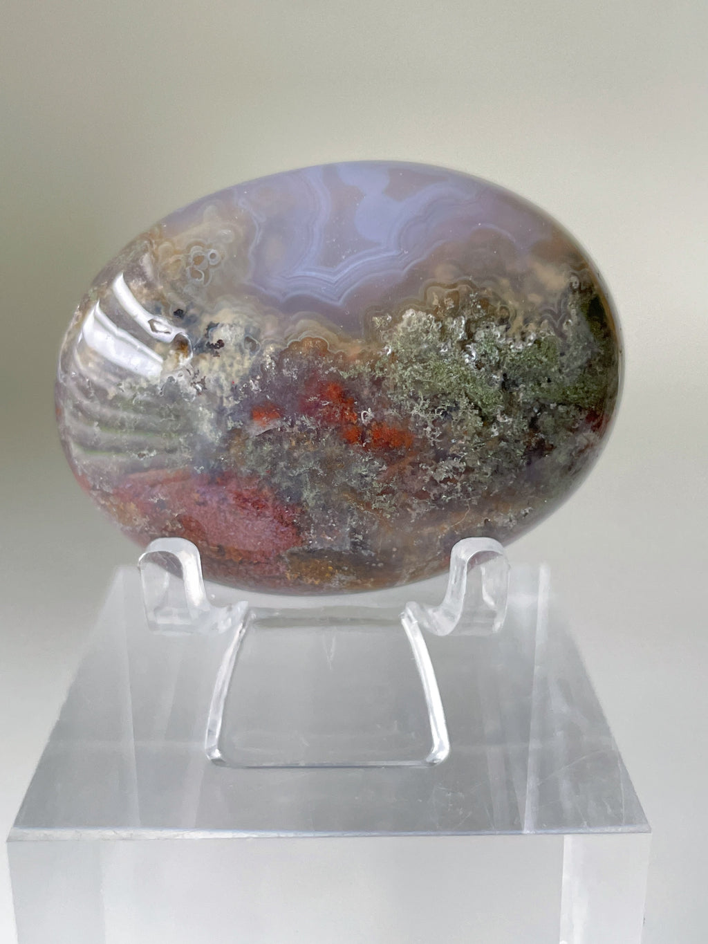 Scenic moss agate palm stone displayed on a clear stand, showcasing earthy tones and intricate patterns.