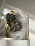 Scenic moss agate tower featuring earthy tones and intricate patterns, displayed on a clear pedestal.