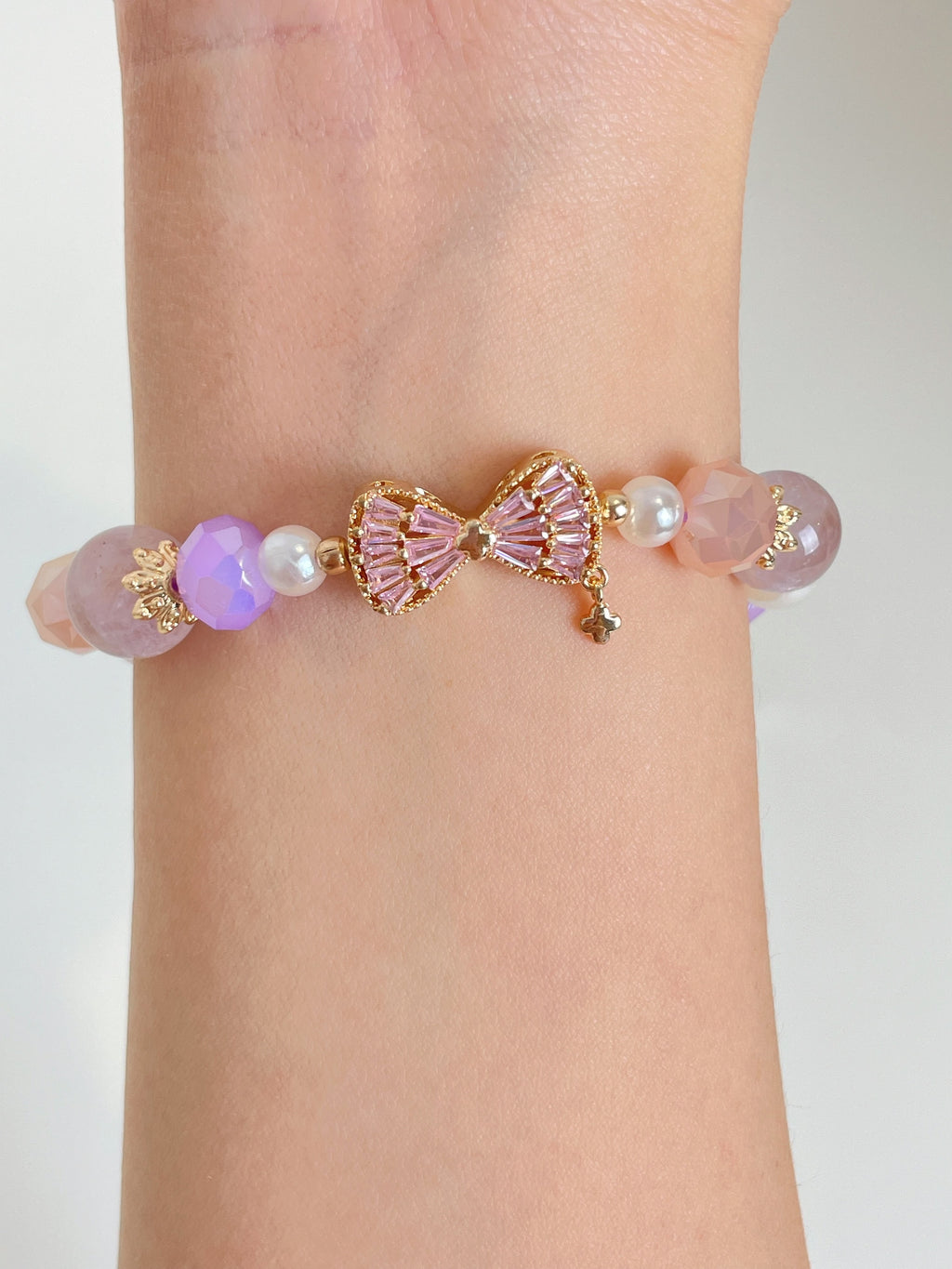 A delicate bracelet featuring pink and purple beads, adorned with a gold bow charm and pearl accents.