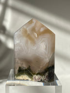 Scenic moss agate crystal tower with soft hues and earthy patterns, displayed on a clear pedestal.