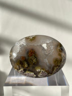 Scenic moss agate palm stone with earthy tones and unique patterns, displayed on a clear stand.